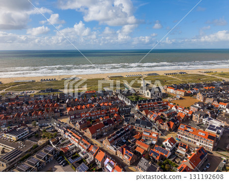 Aerial view of a coastal town with dense red roofed buildings, a tall steepled church, green spaces, wide sandy beach with huts, and ocean waves under a partly cloudy sky. 131192608