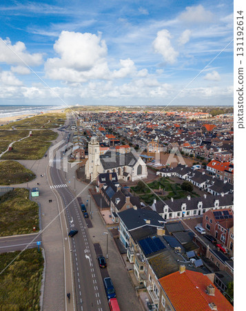Aerial view of a coastal town with red roofed houses, a white church and bell tower near dunes and beach, main road along the shore, and partly cloudy skies adding depth. 131192614
