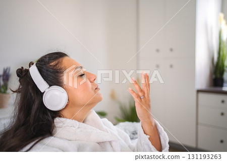 Young woman wearing bathrobe listening to music with headphones at home Young woman wearing bathrobe listening to music with headphones at home 131193263