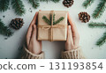 A pair of hands in cozy sweater holding a natural gift box with pine twig decor, surrounded by fir branches and pinecones on white background, Christmas decor and craftwork, eco friendly, sustainable 131193854