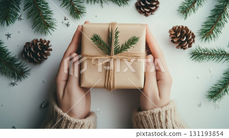 A pair of hands in cozy sweater holding a natural gift box with pine twig decor, surrounded by fir branches and pinecones on white background, Christmas decor and craftwork, eco friendly, sustainable 131193854