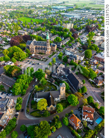 Aerial view of a European town with a tall green spire gothic church at its center, surrounded by red roofed houses, winding streets, trees, and fields blending into rural outskirts. 131197570