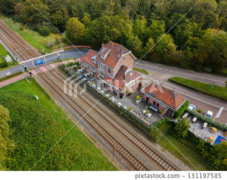 Aerial view of a red roofed cafe beside railway tracks and a road crossing with safety barriers, set in a green semi rural landscape of trees and vegetation. Aerial view of a red roofed cafe beside railway tracks and a road crossing with safety barriers, set in a green semi rural landscape of trees and vegetation. 131197585
