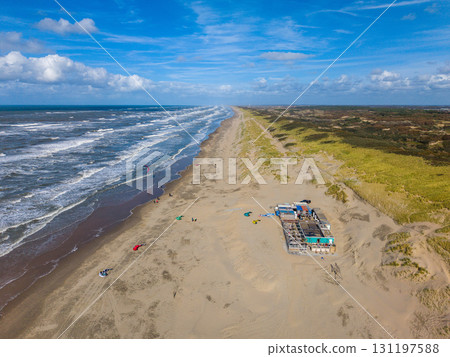 Aerial view of a sandy beach bordered by vegetated dunes, with a small building nearby, people on the shore, and colorful kites hinting at kite surfing under a partly cloudy sky. 131197588