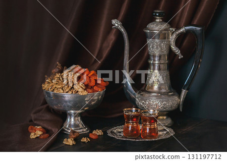 Still life with Ancient East metallic crockery - vintage jug, vase with dried fruit and plate with tea in armuda glasses 131197712