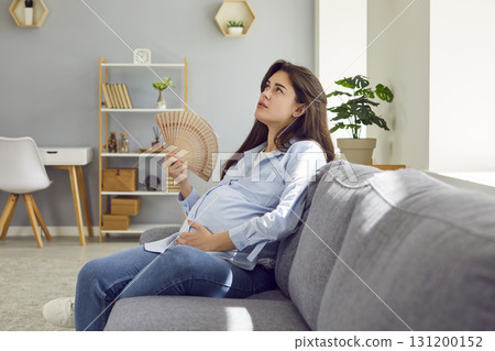 Young expectant mother, tired of summer heat, sitting on sofa and cooling herself with hand fan 131200152
