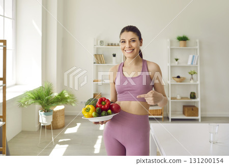 Woman in sportswear standing at home after working out with a plate of vegetables. Woman in sportswear standing at home after working out with a plate of vegetables. 131200154
