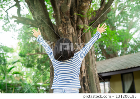 Happy woman with open arms embracing nature under large tree in sunny garden. Natural healing therapy. Social media detox retreat at eco hotel. Nature connection wellness at sustainable eco retreat. 131200183