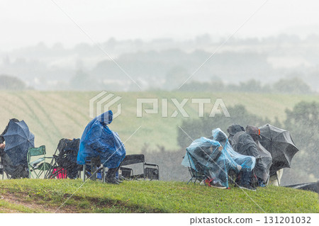 Spectators sit on chairs under heavy rain with umbrellas and rain ponchos, showing resilience and dedication at sport event. Wet weather countryside fans continue follow outdoor sporting tournament 131201032