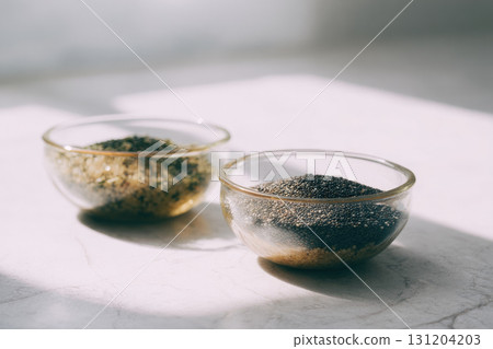 Glass bowls filled with chia seeds and quinoa on white marble surface, sunlight casting soft shadows, healthy food concept, minimal style, natural light 131204203
