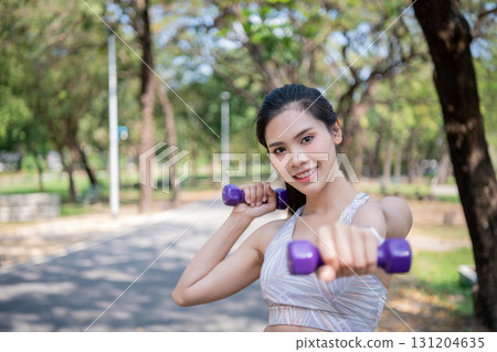 A young woman joyfully exercising outdoors with dumbbells in a sunny park. 131204635
