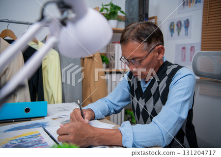 An elderly man in a sweater vest diligently sketches designs at his workspace. 131204717