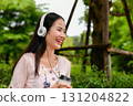 A young woman enjoys her coffee while wearing headphones outdoors, radiating joy in a serene green environment. 131204822