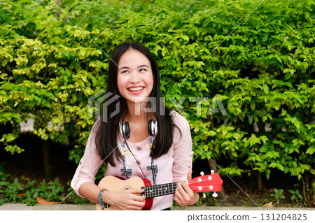 Joyful girl playing ukulele outdoors, radiating happiness in a lush green environment. Joyful girl playing ukulele outdoors, radiating happiness in a lush green environment. 131204825