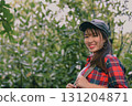Cheerful young woman enjoying nature, wearing a cap and plaid shirt, smiling amidst lush greenery. 131204871