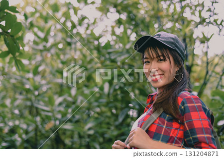 Cheerful young woman enjoying nature, wearing a cap and plaid shirt, smiling amidst lush greenery. Cheerful young woman enjoying nature, wearing a cap and plaid shirt, smiling amidst lush greenery. 131204871