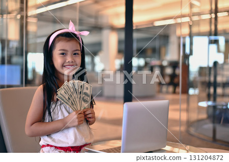 Young girl proudly holds cash near a laptop, representing financial success and ambition in the modern world. Young girl proudly holds cash near a laptop, representing financial success and ambition in the modern world. 131204872