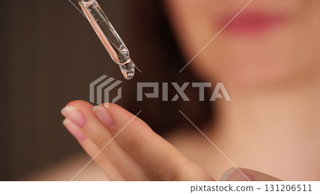 Young woman holding a dropper with serum for face. Facial oil fall from a glass pipette, macro view. Selective focus. Cosmetic background with liquid skincare product. Beauty treatment concept. Young woman holding a dropper with serum for face. Facial oil fall from a glass pipette, macro view. Selective focus. Cosmetic background with liquid skincare product. Beauty treatment concept. 131206511