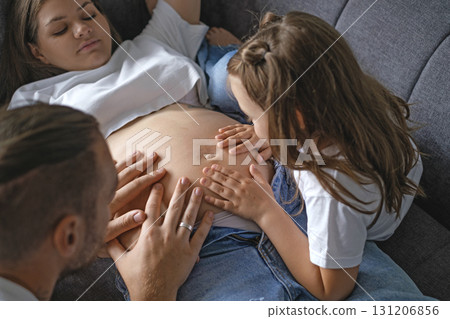 A mother pregnant with second child is lying on the couch with her youngest daughter and her husband, surrounded by love and care. The father touches the belly and feels the baby's thrusts in womb A mother pregnant with second child is lying on the couch with her youngest daughter and her husband, surrounded by love and care. The father touches the belly and feels the baby's thrusts in womb 131206856