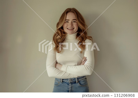 A cheerful young woman with wavy hair stands confidently with arms crossed, wearing a light sweater and jeans in a simple setting 131206908