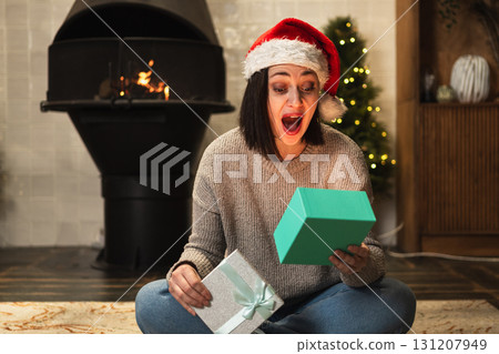 Merry Christmas. Young woman in Santa hat unpacking open up gift box near Christmas tree. Girl in living room with Christmas tree, fireplace opening gift box with surprise face. Christmas eve at home Merry Christmas. Young woman in Santa hat unpacking open up gift box near Christmas tree. Girl in living room with Christmas tree, fireplace opening gift box with surprise face. Christmas eve at home 131207949