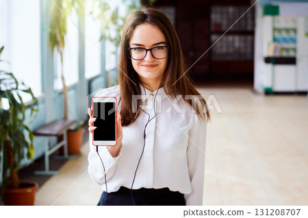 Portrait of a happy cheerful girl student with headphones holding black screen mobile phone and smiling to the camera over background of college corridor Portrait of a happy cheerful girl student with headphones holding black screen mobile phone and smiling to the camera over background of college corridor 131208707