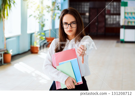 Front view of sad and critique student girl showing thumbs down, looking to the camera and holding books, pages, exercise books. Concept of boring uninteresting learning, studying 131208712