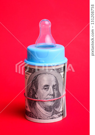 Nipple of baby milk bottle on a roll of dollar cash against red background. Concept of high price of bringing up Nipple of baby milk bottle on a roll of dollar cash against red background. Concept of high price of bringing up 131208758