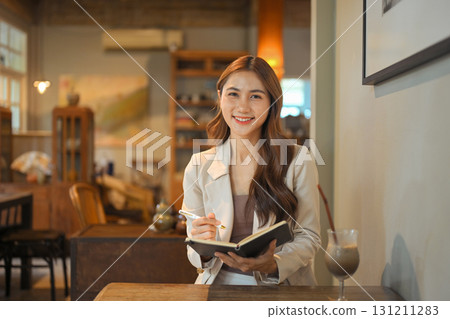 Smiling businesswoman holding a notebook and pen while sitting in a cozy cafe 131211283