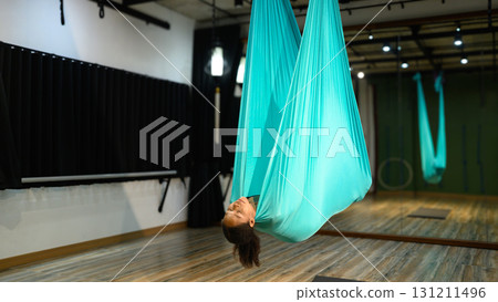 Young woman practicing aerial yoga inside turquoise silk hammock in fitness studio 131211496
