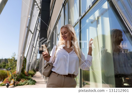 Young woman checks her phone while leaning against a modern building on a sunny day Young woman checks her phone while leaning against a modern building on a sunny day 131212901