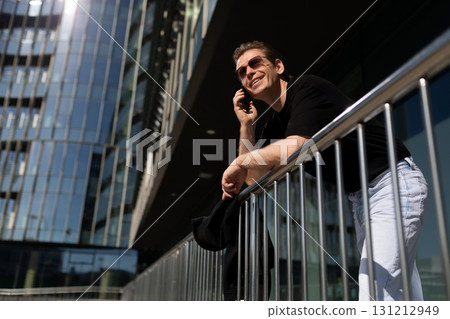 Man talking on the phone while leaning on a railing outside a modern building on a sunny day Man talking on the phone while leaning on a railing outside a modern building on a sunny day 131212949