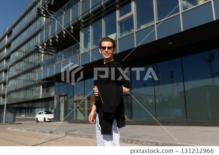 Young man enjoys a sunny day outside a modern building while casually holding a jacket Young man enjoys a sunny day outside a modern building while casually holding a jacket 131212966