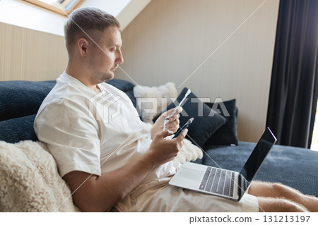 Man working remotely at home with laptop and smartphone, making online payment with credit card. Concept of digital banking, fintech, e-commerce, modern lifestyle and financial management. 131213197