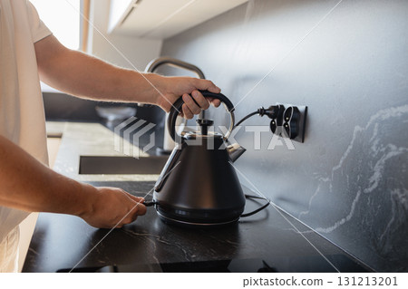 Man placing black electric kettle on modern kitchen stove Man placing black electric kettle on modern kitchen stove 131213201