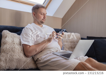 Man working on laptop and using smartphone while sitting on sofa at home. Concept of freelancing, digital lifestyle, mobile technology, e-banking, online shopping, communication and productivity. Man working on laptop and using smartphone while sitting on sofa at home. Concept of freelancing, digital lifestyle, mobile technology, e-banking, online shopping, communication and productivity. 131213216
