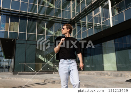 Man walking confidently in modern urban setting wearing stylish outfit with sunglasses and carrying 131213439