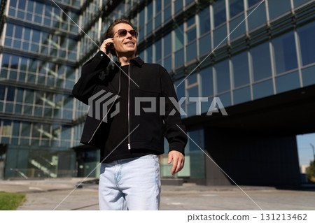 Young man wearing sunglasses talks on a phone outside modern building during bright sunny day in Young man wearing sunglasses talks on a phone outside modern building during bright sunny day in 131213462