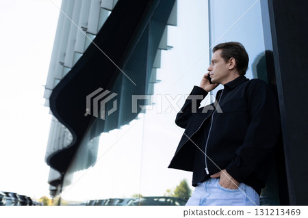 Man talking on the phone while leaning against a modern building in an urban area during daylight 131213469