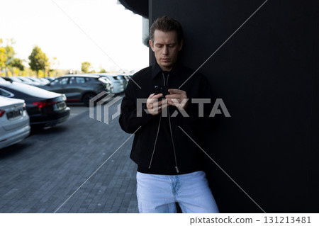 Casual man using smartphone while leaning against a wall in a parking lot during daylight hours Casual man using smartphone while leaning against a wall in a parking lot during daylight hours 131213481