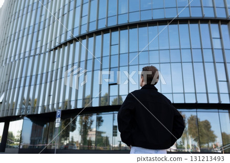 Young man admires modern architecture of glass building in urban setting during daytime stroll 131213493