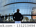 Man stands in front of large modern building reflecting blue sky and surrounding trees at midday 131213494