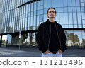 Man standing confidently in front of modern glass building during daylight in an urban setting 131213496
