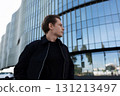 Young man in black jacket standing near modern glass building in urban setting during late afternoon 131213497