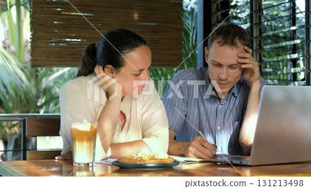 On terrace of coffee shop a man and woman discuss a new project using a laptop and taking notes with coffee on the table  131213498