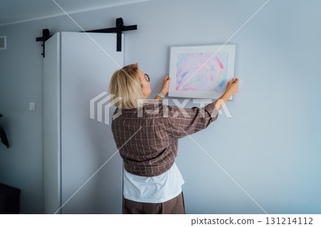 Side view middle aged woman putting painting picture in frame on the wall. Casual lady taking care of coziness in her new stylish apartment. Mature female decorating room. Moving and renovate house Side view middle aged woman putting painting picture in frame on the wall. Casual lady taking care of coziness in her new stylish apartment. Mature female decorating room. Moving and renovate house 131214112