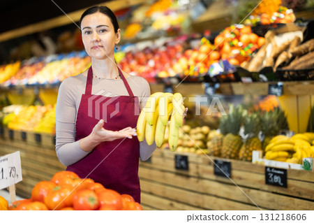 Positive female seller offers to buy ripe tasty bananas in grocery department of supermarket 131218606