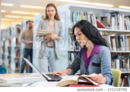 Asian woman student in the library, searches for information in books and types it in a laptop Asian woman student in the library, searches for information in books and types it in a laptop 131218798