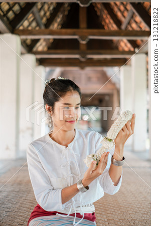 A woman in traditional Thai attire holds a jasmine garland, showcasing cultural heritage and elegance in a serene architectural setting. A woman in traditional Thai attire holds a jasmine garland, showcasing cultural heritage and elegance in a serene architectural setting. 131218822