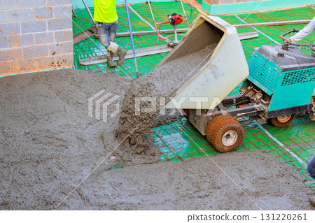 Workers are busy pouring concrete onto floor at construction site, uses wheeled concrete wheelbarrow during build. 131220261
